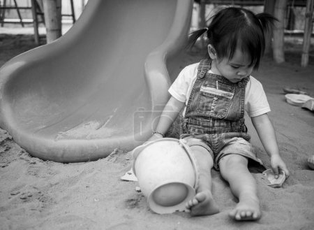 Photo for "Asian little girl sitting on sand ground in the playground and play a sand with plastic beach toys happily. Playing is learning for children. Black-white picture." - Royalty Free Image