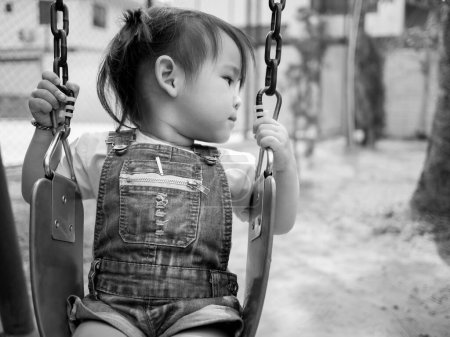 Téléchargez les photos : "Asiatique petit enfant est assis sur une balançoire heureusement dans la cour de récréation. Jouer, c'est apprendre pour les enfants. Photo noir-blanc." - en image libre de droit