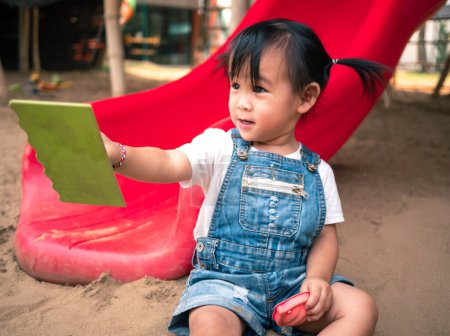 Téléchargez les photos : Asiatique petite fille assis sur le sol de sable dans la cour de récréation et jouer un sable avec des jouets de plage en plastique heureux. - en image libre de droit