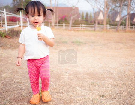 Photo for "Asian little girl walking and eating ice cream deliciously in summer in the park at Chiang mai, Thailand." - Royalty Free Image