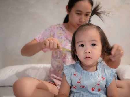 Foto de Encantadora niña asiática está mirando a la cámara y sonriendo mientras su joven madre está peinando el cabello de su hija por la mañana en la habitación - Imagen libre de derechos