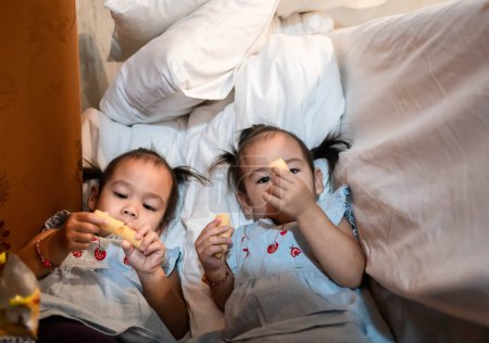Foto de Asiática niña con hermana comiendo bocadillo deliciosamente juntos en el dormitorio thr - Imagen libre de derechos