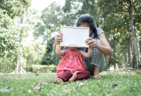 Photo for Asian little girl holding empty white board with mom and sitting on grasses ground. Space for write your text - Royalty Free Image