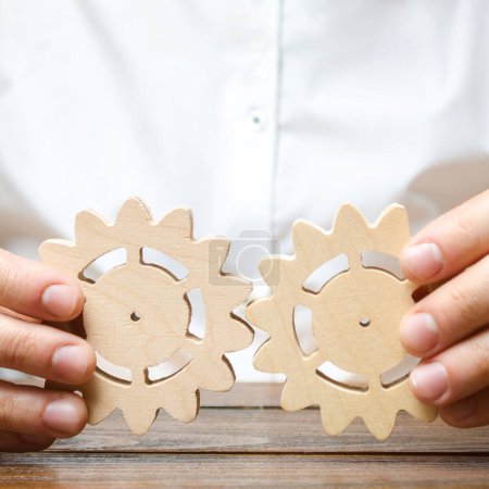 Photo for "Businessman in white shirt connects two wooden gears. Improving work efficiency, establishing new connections and suppliers. Symbolism of establishing business processes and communication." - Royalty Free Image