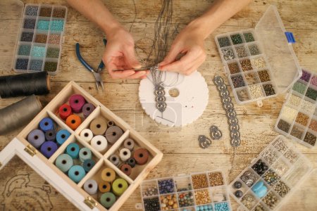 Draufsicht weiblicher Hände bei der Herstellung eines Makrambands mit Kumihimo-Scheibe auf Holztisch mit Werkzeugen, Garnspulen, Natursteinen und farbigen Perlen.