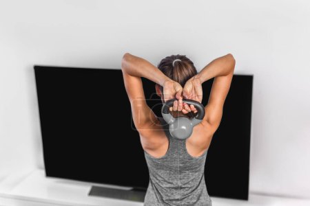 Photo for "Home exercise strength training workout overhead triceps extension muscles exercises woman watching online tv fitness class live streaming at home. Gym girl lifting kettlebell weights" - Royalty Free Image
