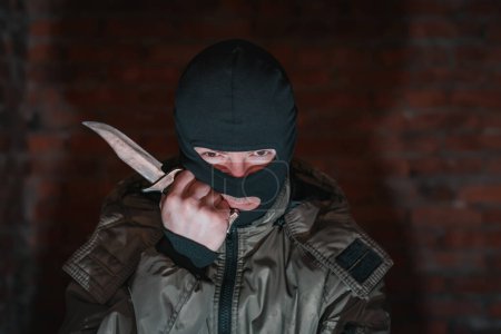 Téléchargez les photos : Bandit fatigué dans la cagoule et tenant un couteau sur un fond de mur de briques - en image libre de droit