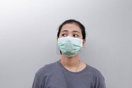 Foto de Mujer joven de Asia que usa mascarilla médica mientras está de pie contra la pared gris - Imagen libre de derechos