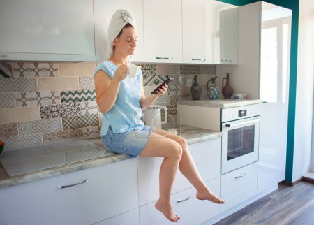 Téléchargez les photos : Femme avec une serviette sur la tête dans sa cuisine prendre le petit déjeuner et regarder le téléphone - en image libre de droit