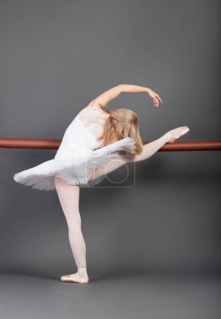 Foto de Joven bailarina de ballet estirándose en la barra de ballet sobre fondo gris - Imagen libre de derechos