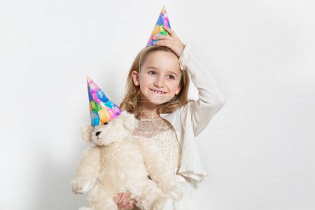 Foto de Feliz joven cumpleañera con sombrero de fiesta con oso de peluche sobre fondo de color - Imagen libre de derechos