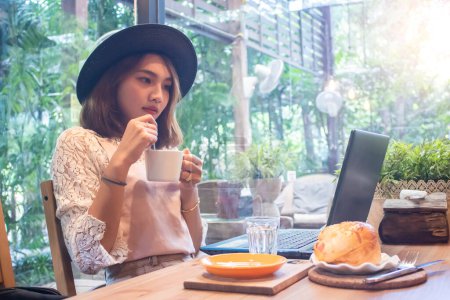 Téléchargez les photos : Femme asiatique tenant tasse de café regardant ordinateur portable dans le café - en image libre de droit