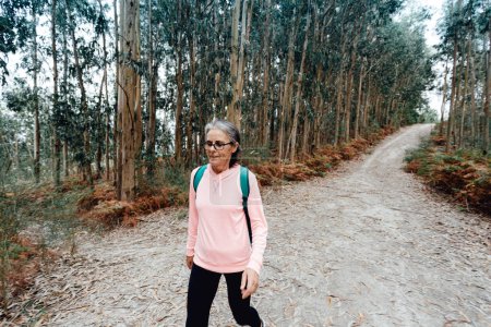 Photo for Old woman walking in the forest with sport clothes, bag and glasses while smiling - Royalty Free Image