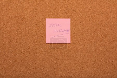 Foto de Recordatorio Social distanciamiento escrito a mano en una pegatina rosa en un tablón de anuncios de corcho. - Imagen libre de derechos