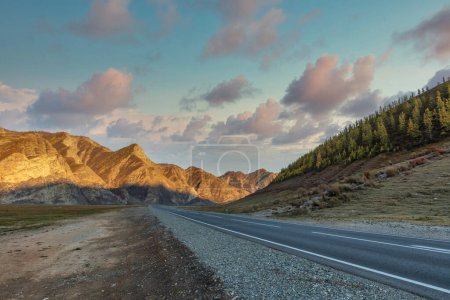 Téléchargez les photos : Vue panoramique à angle bas de la crête de montagne. Autoroute au premier plan menant jusqu'aux montagnes. Beau ciel nuageux au coucher du soleil comme toile de fond. L'heure dorée. Montagnes Altaï, Sibérie, Russie - en image libre de droit