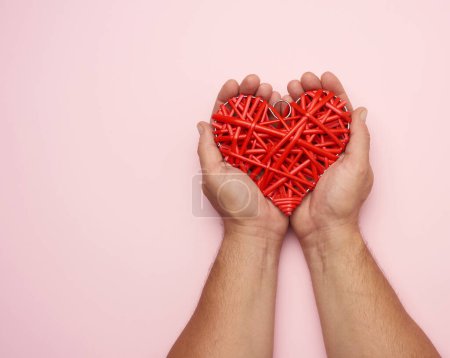 Téléchargez les photos : Deux mains masculines tenant un cœur en osier rouge sur un fond rose - en image libre de droit