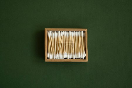 Foto de Hisopos de algodón ecológicos. Una caja de cartón que contiene cogollos de algodón de bambú reciclables sobre una superficie verde. Concepto de cero residuos. Producto ecológico. Palos cosméticos en una caja. Palos de madera para limpiar las orejas - Imagen libre de derechos