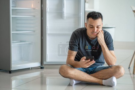 Foto de "Un joven pide comida con un smartphone. Refrigerador vacío sin comida. Servicio de entrega de alimentos publicidad" - Imagen libre de derechos