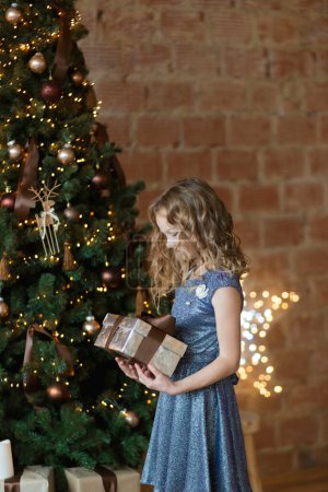 Foto de La adorable niña en vestido precioso sostiene caja de regalo de pie cerca del árbol - Imagen libre de derechos