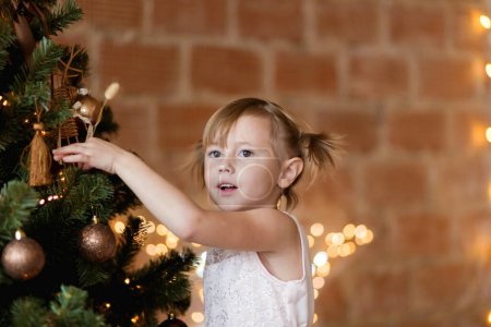 Foto de Linda niña en un vestido elegante cuelga juguetes de Navidad en el árbol - Imagen libre de derechos