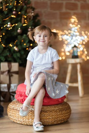 Foto de Pretty little child is sitting in front of christmas tree among garlands - Imagen libre de derechos