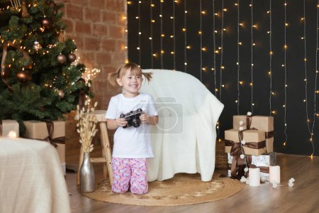 Téléchargez les photos : Adorable petite fille en pyjama assis sur le sol avec caméra dans ses mains - en image libre de droit