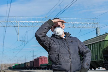 Téléchargez les photos : Un contremaître portant un masque médical dans une gare surveille les trains, se couvrant le visage avec sa main du soleil - en image libre de droit