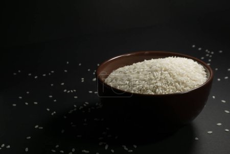 Photo for White uncooked rice in a bowl with scattered grains nearby on a black background - Royalty Free Image