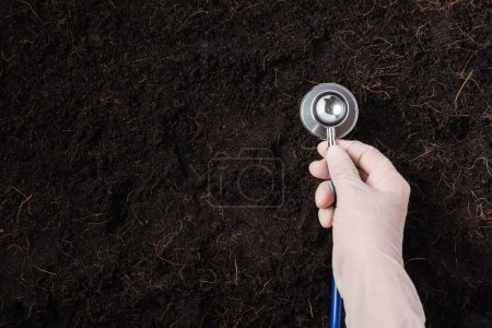 Photo for Hand of researcher holding a stethoscope on fertile black soil - Royalty Free Image