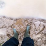 Feet in green rubber boots stands in wet brown mud puddle directly above view