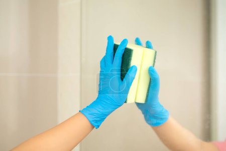 Téléchargez les photos : Gros plan femmes main dans un gant en caoutchouc bleu et brosse de nettoyage dans la photo, enlève et lave lavabo de salle de bains et miroir. - en image libre de droit