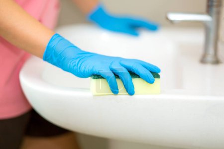 Téléchargez les photos : Une main dans un gant de caoutchouc bleu dans l'image, enlève et lave lavabo de salle de bains - en image libre de droit