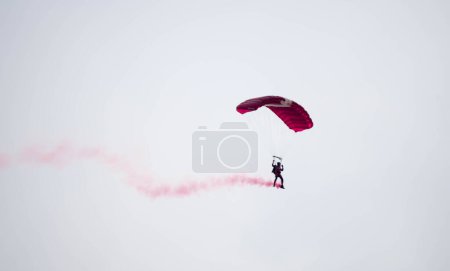 Téléchargez les photos : Silhouette parachute cascade floue et floue tout en glissant dans les airs avec traînée de fumée rouge lors d'une exposition aérienne - en image libre de droit