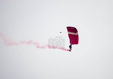 Téléchargez les photos : Silhouette parachute cascade floue et floue tout en glissant dans les airs avec traînée de fumée rouge lors d'une exposition aérienne - en image libre de droit