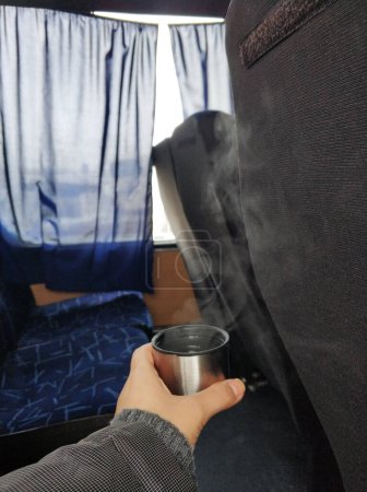 Foto de Un hombre sostiene en su mano una gorra de un termo con té caliente humeante, en la cabina del autobús. Bebida caliente durante el viaje - Imagen libre de derechos
