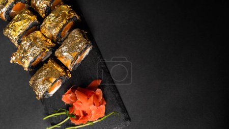 Foto de Rollos de sushi personalizados con nori, tinta de sepia, salmón fresco, queso crema, calabaza de pimienta, anguila, salsa unagi y pan de oro comestible con jengibre rojo, wasabi en un plato cuadrado sobre mesa negra y fondo - Imagen libre de derechos