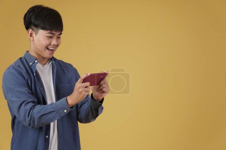 Photo for "portrait of excited young asian man dressed casually playing video games on smartphone isolated on yellow background" - Royalty Free Image