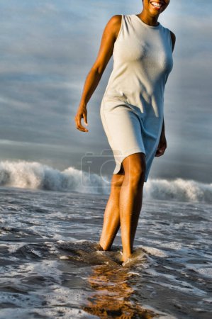 Téléchargez les photos : "Femme afro-américaine joyeuse marchant à travers le surf sur la plage" - en image libre de droit
