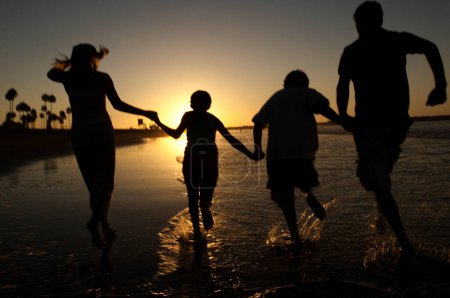 Téléchargez les photos : Vue arrière de la famille heureuse profitant du coucher de soleil sur la plage - en image libre de droit