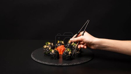 Photo for Hand with chopsticks wants to take custom sushi roll with black rice, crab meat, avocado, smoked salmon mousse, oar caviar, masago, shrimp cocktail, edible gold leaf, ginger, wasabi on black table. - Royalty Free Image