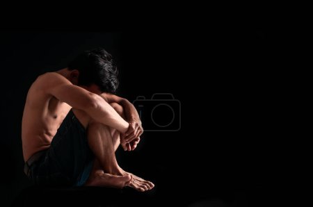 Téléchargez les photos : Triste homme étreint son genou avec la dépression assis seul sur le sol dans la chambre noire - en image libre de droit