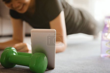Photo for "A sporty woman using smartphone during workout at home in the living room. Online personal trainer or on mobile phone.Sport and recreation concept." - Royalty Free Image