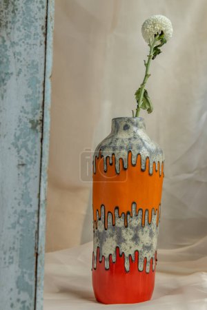 Téléchargez les photos : "Fleur blanche en vase en céramique à la main orange et rouge sur nappe texturée Blush. " - en image libre de droit