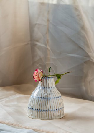 Foto de "Flor rosa sobre jarrón de cerámica hecho a mano de color cobre y sobre mantel texturizado rubor. " - Imagen libre de derechos