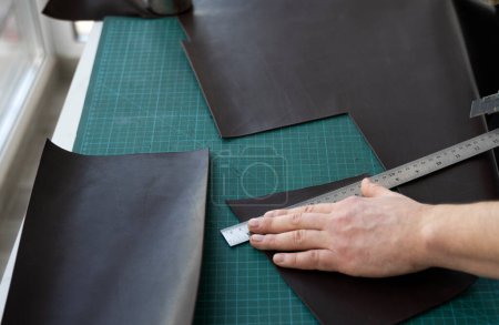 Photo for "Men s hand holding a stationery knife and metal ruler and cutting on a pieces for a leather wallet in his workshop. Working process with a brown natural leather. Craftsman holding a crafting tools." - Royalty Free Image