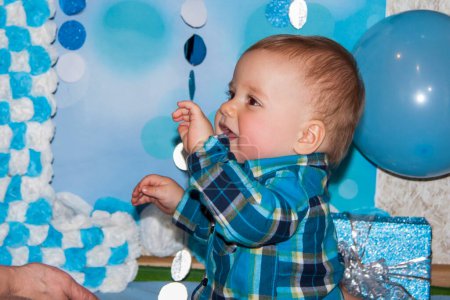 Téléchargez les photos : Un beau bébé fête son premier anniversaire. Portrait du bébé. Un garçon en chemise bleue parmi les ballons et les cadeaux. - en image libre de droit
