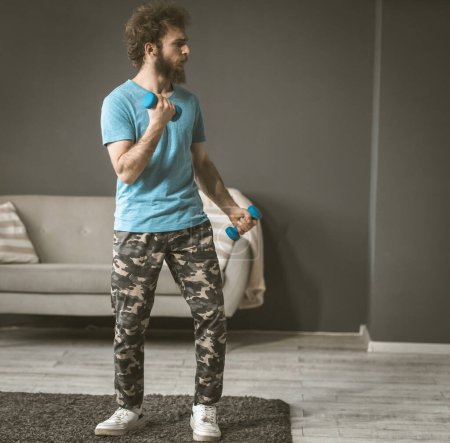 Photo for "A young man sports in his room. Exercise with dumbbells. The curly-haired guy is holding dumbbells in his hands." - Royalty Free Image