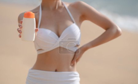 Foto de "Mujer en bikini blanco sosteniendo botella de protector solar en la mano en la playa." - Imagen libre de derechos