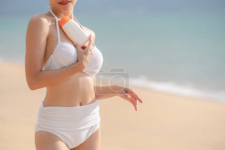 Foto de Mujer en bikini blanco sosteniendo botella de protector solar en la mano en la playa. - Imagen libre de derechos