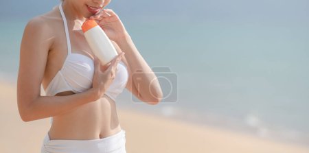 Téléchargez les photos : Femme en bikini blanc tenant bouteille de crème solaire à la main sur la plage - en image libre de droit